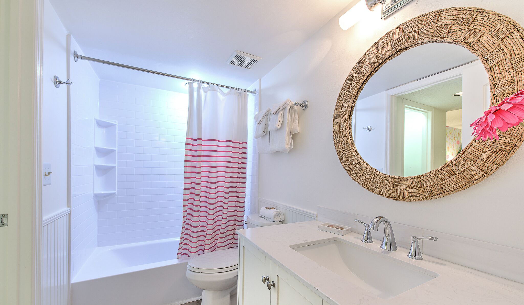 Een badkamer met een bad, een toilet, een wastafel met twee kraan en een spiegel met geweven frame, plus een douchegordijn met rode strepen.