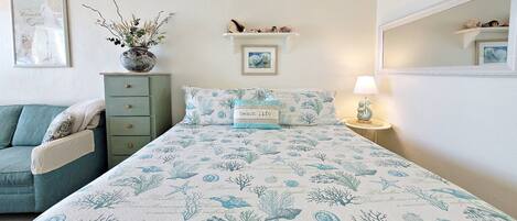 A bedroom with a bed, a dresser, a nightstand, a lamp, and a wall-mounted shelf with decorative items.
