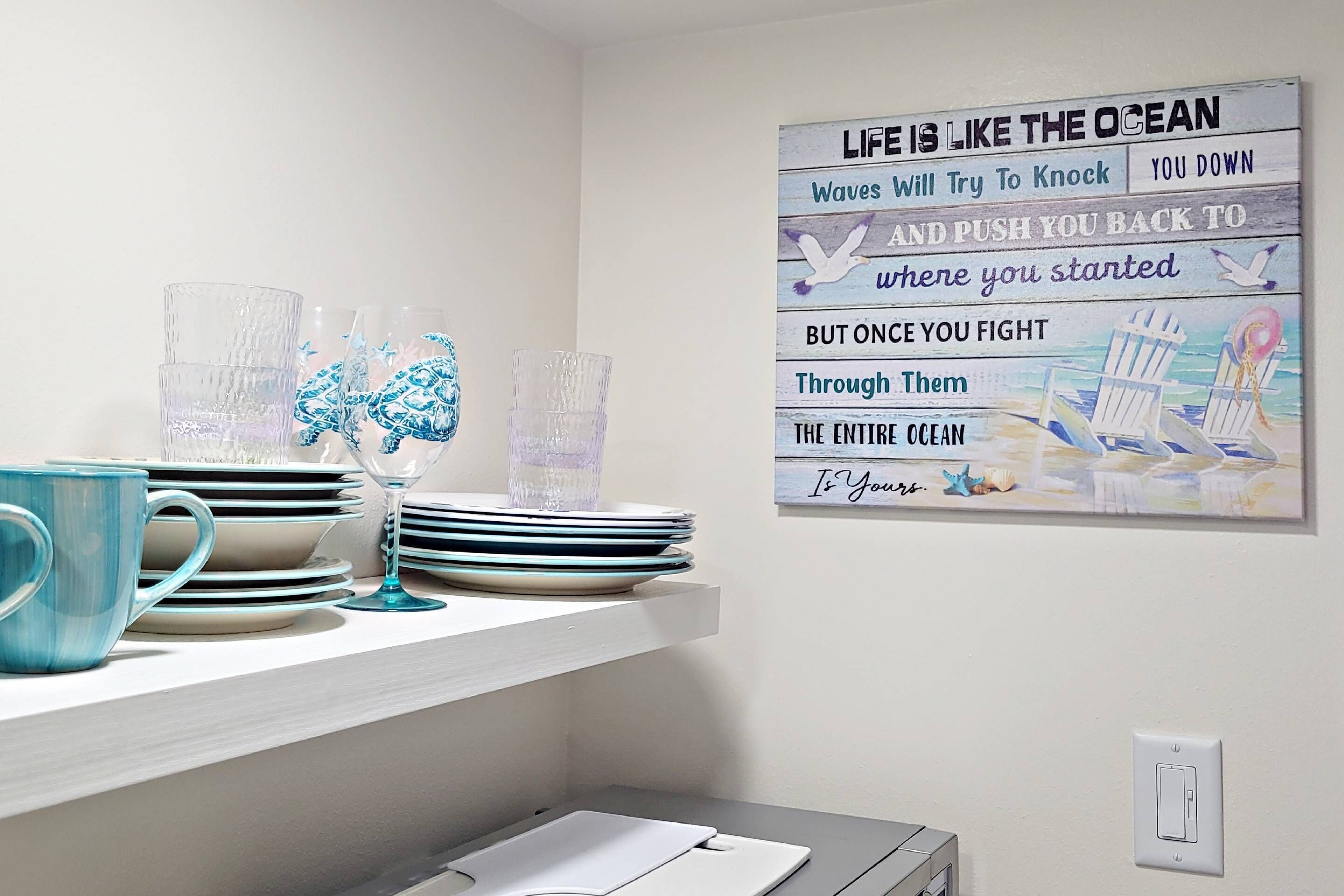 Una cocina con encimera blanca, estante de pared con platos, vasos y una taza azul. Una cita motivacional está expuesta en la pared.
