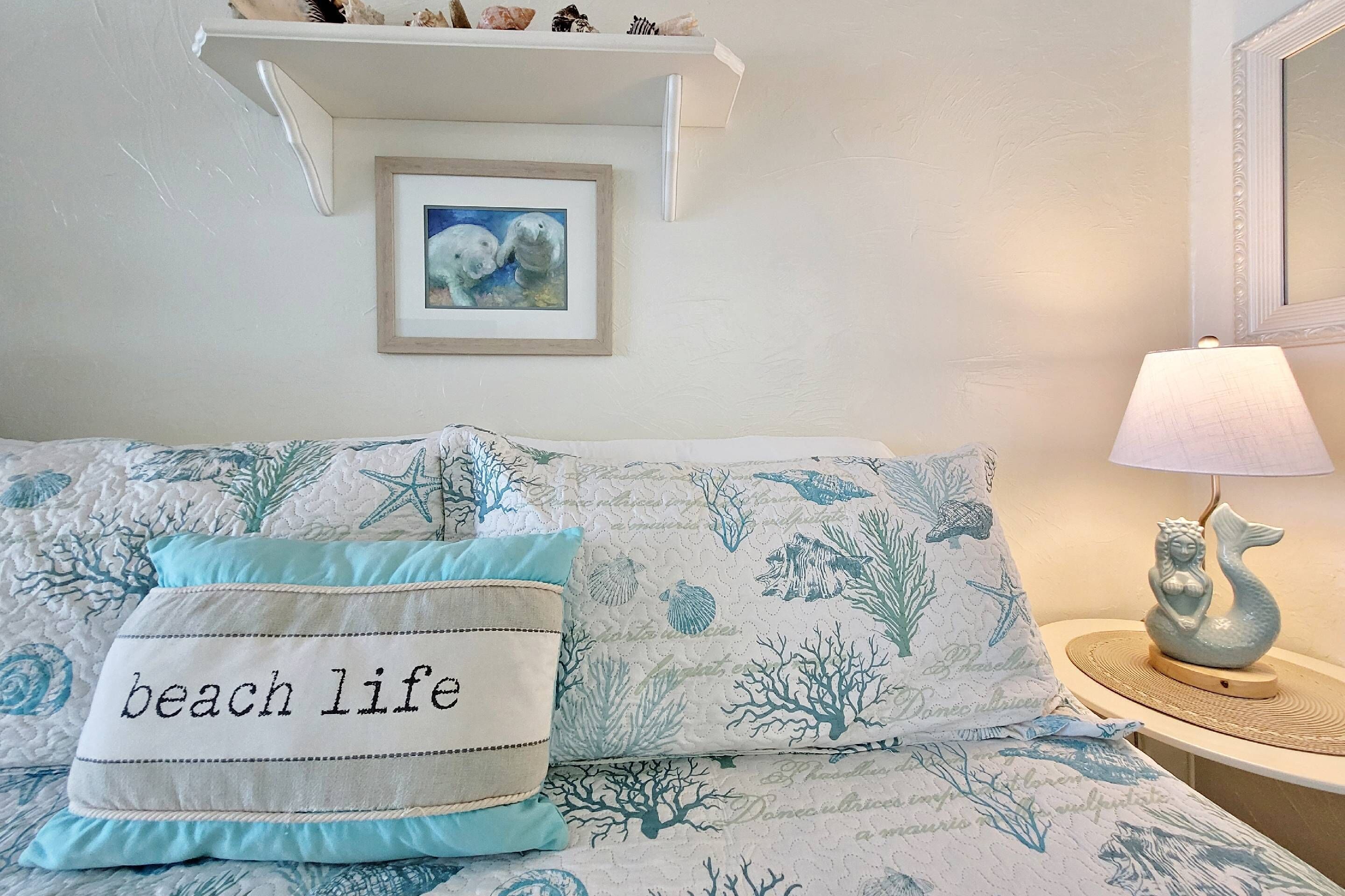 Ein Schlafzimmer mit einem Bett, das mit einem Küstenmotiv bezogen ist, sowie mit passenden Kissen, einer kleinen Ablage mit Dekoration und einem gerahmten Bild an der Wand.