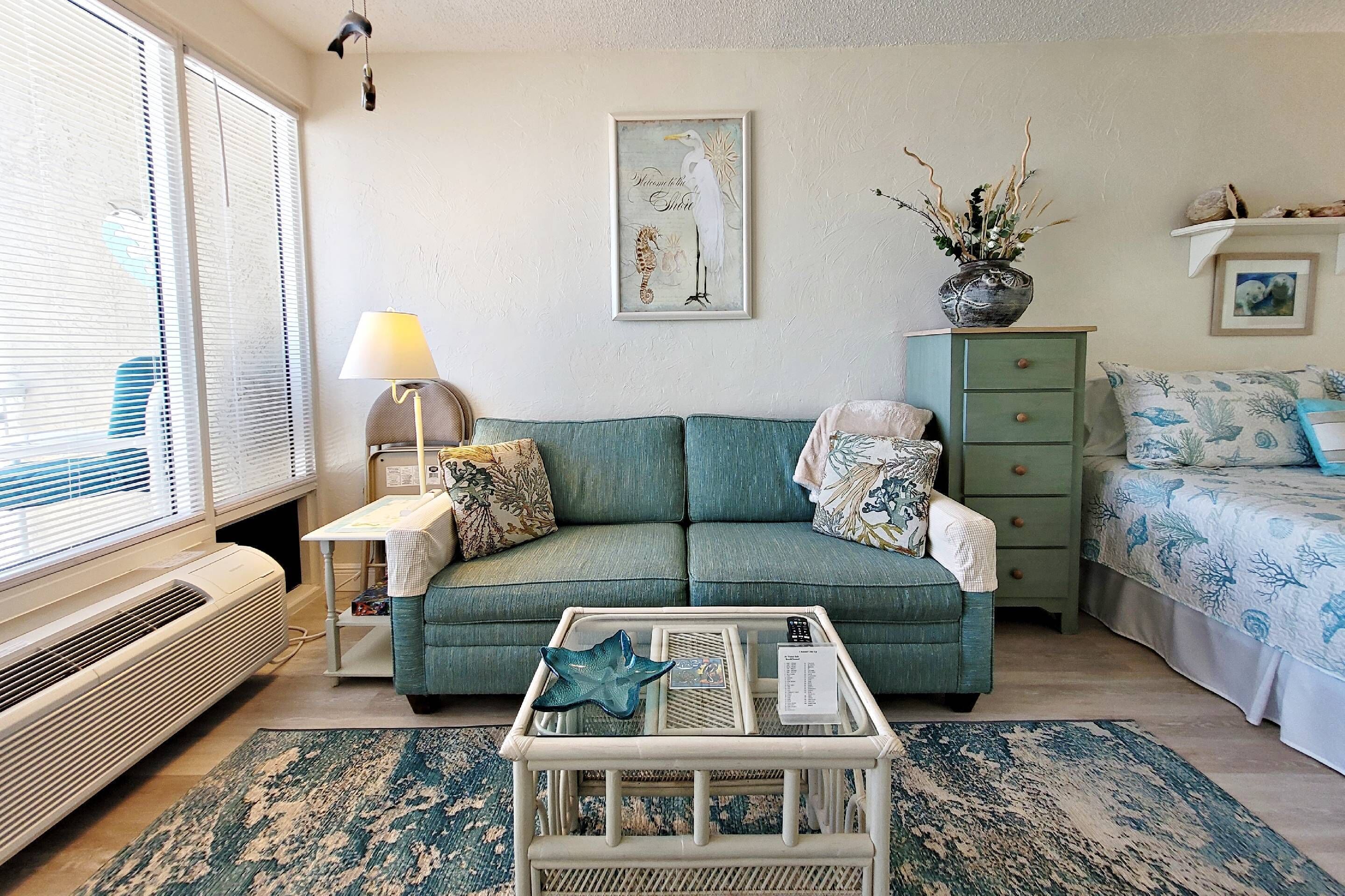A living room with a teal sofa, a white coffee table, a lamp, a framed artwork, a green dresser, and a bed with a floral bedspread.