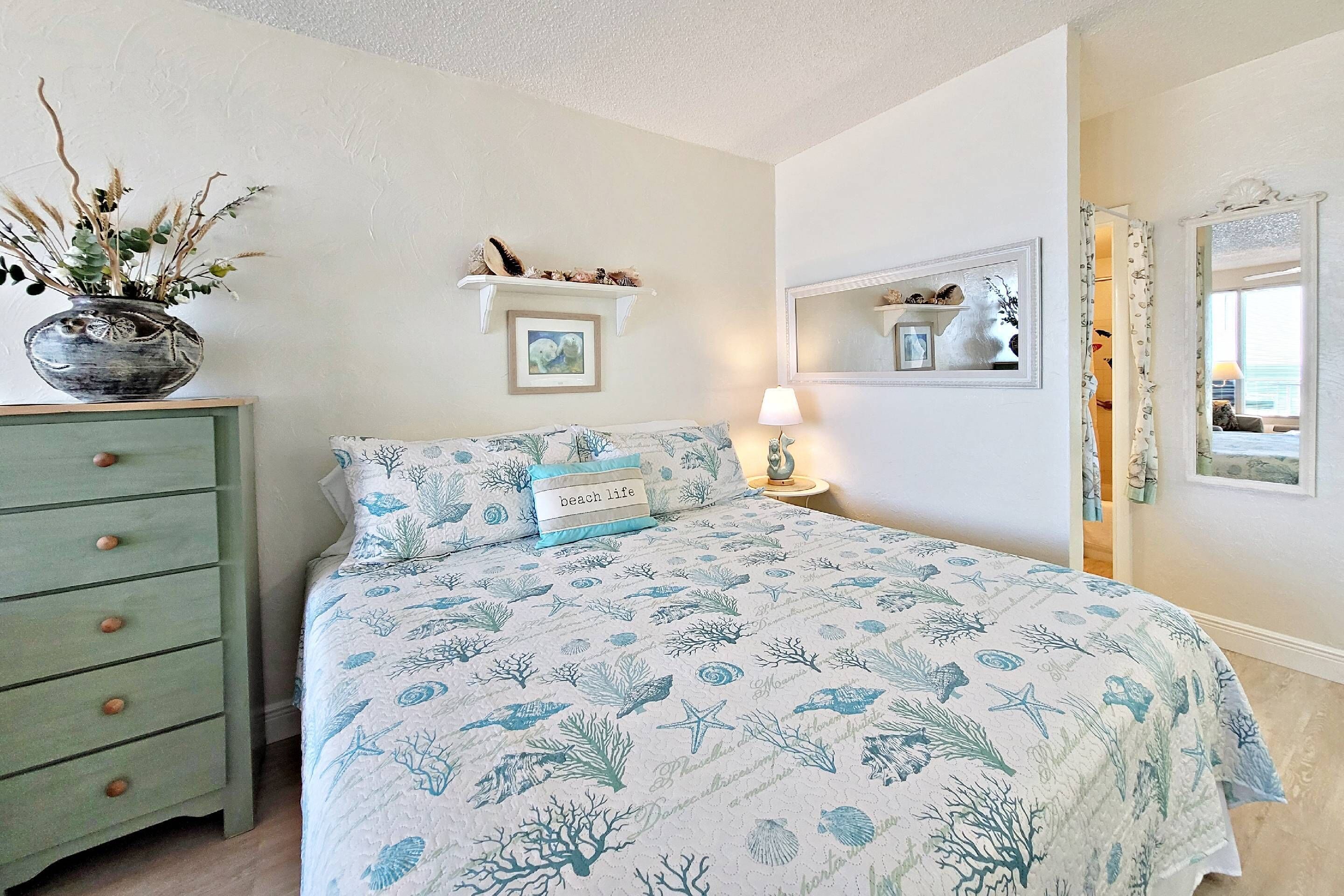 A bedroom with a bed, a dresser, a nightstand, a lamp, a mirror, and a shelf with decorative items.