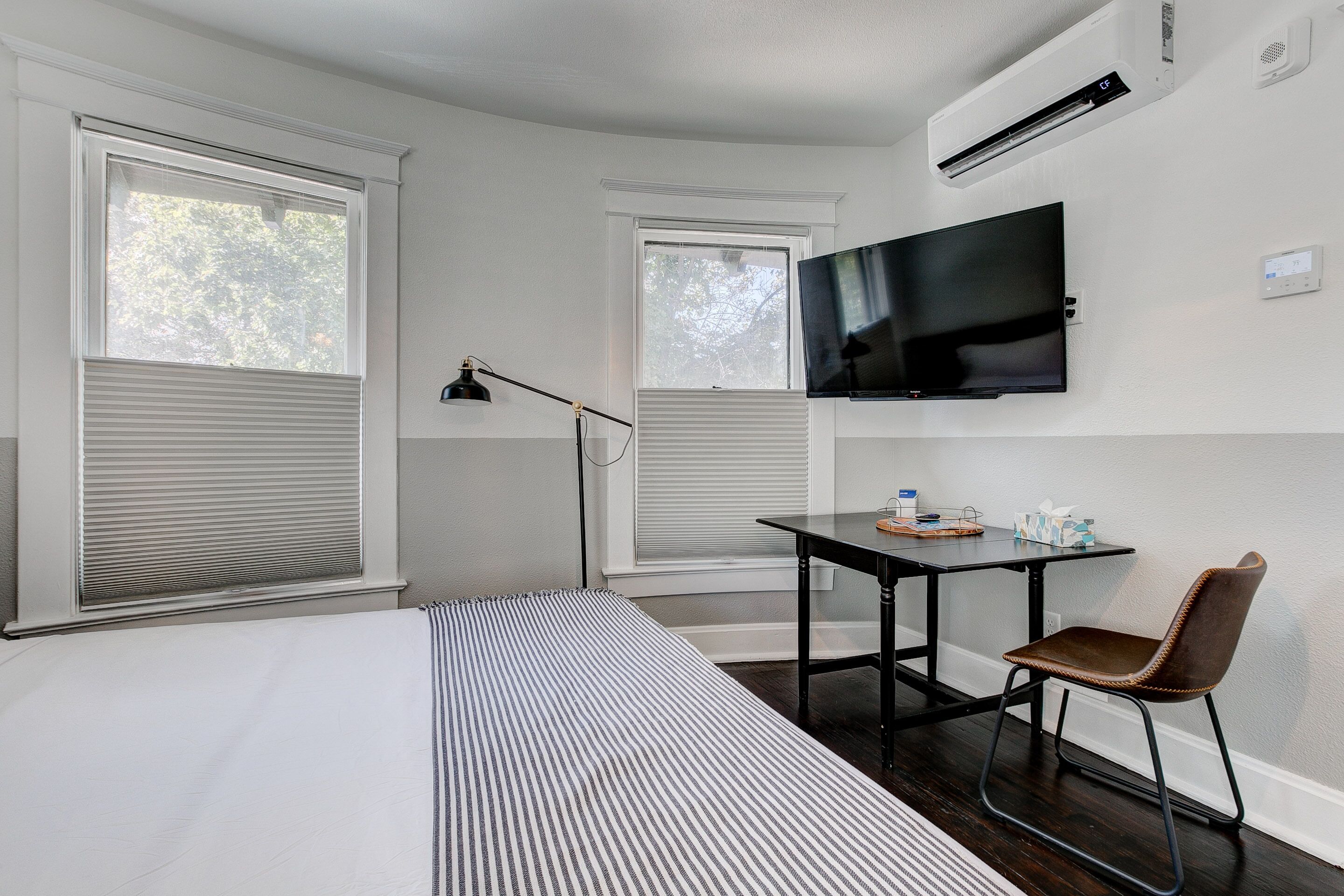 A room with a bed, a desk with a chair, a flat-screen TV, and two windows with blinds.