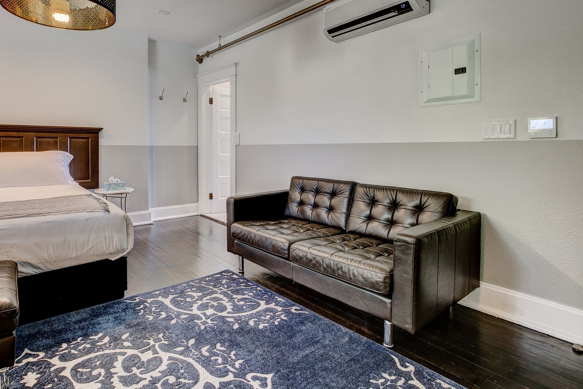 A bedroom with a bed, a leather sofa, a patterned rug, and a wall-mounted air conditioner.