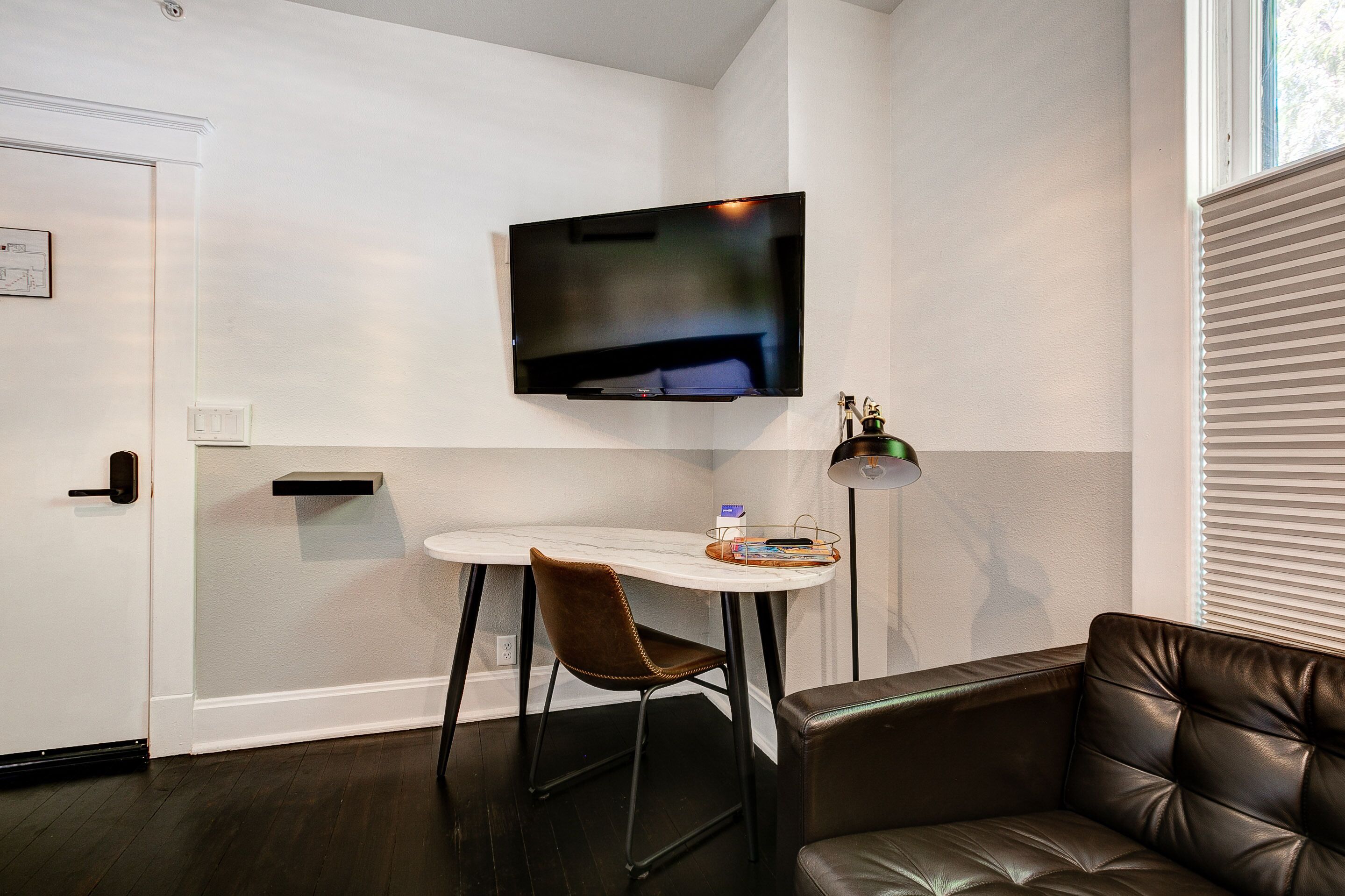 Une chambre moderne dotée d’un téléviseur à écran plat fixé au mur, d’une petite table avec une chaise, d’un canapé en cuir et d’un lampadaire.
