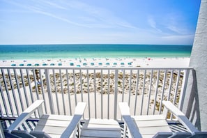 A balcony with white railings overlooking a beach with several blue umbrellas and white lounge chairs.