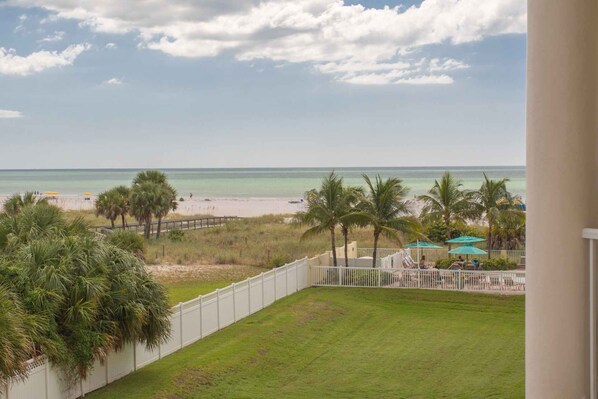A view of a beach with a white fence, palm trees, and a grassy area. - Beachfront Resort.  Balcony with Partial Beach View.  Pools, Cafe, Bar, Fitness Center & Parking (Treasure Island)