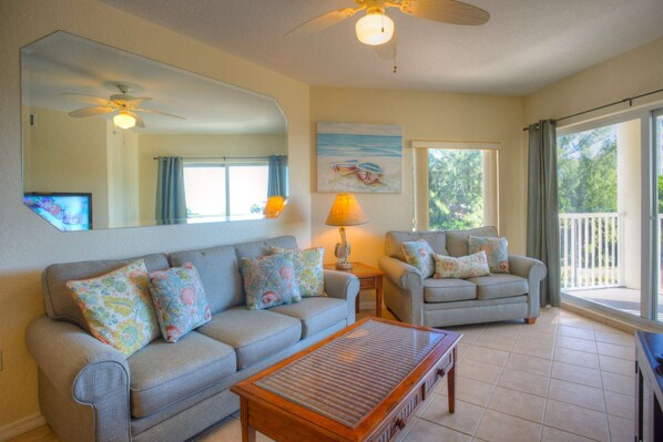 A living room with a sofa, armchair, and a coffee table. There is a ceiling fan, a lamp, and a painting on the wall. - Beachfront Resort.  Balcony with Partial Beach View.  Pools, Cafe, Bar, Fitness Center & Parking (Treasure Island)