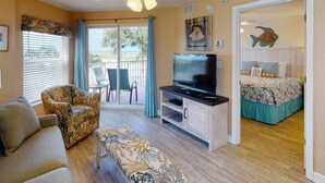 A room with a ceiling fan, a TV on a wooden stand, a sofa, a bench, a bed with a beach-themed bedspread, and a view of the outdoors through a sliding door.