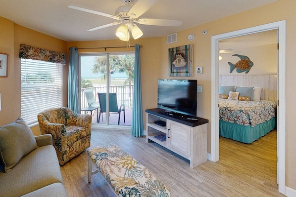 A room with a ceiling fan, a TV on a wooden stand, a sofa, a bench, a bed with a beach-themed bedspread, and a view of the outdoors through a sliding door.