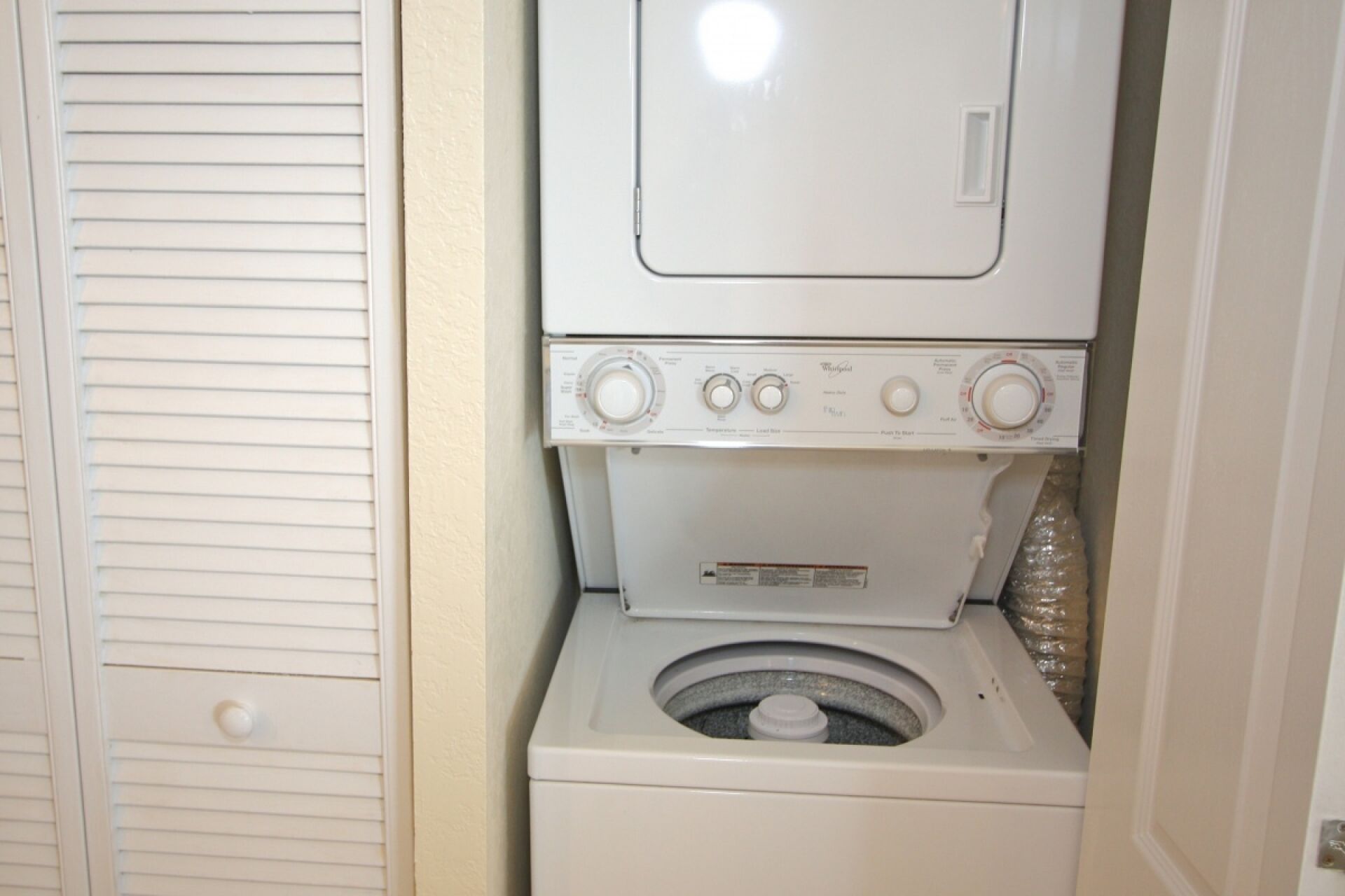 A compact, white, stacked washer and dryer unit in a small laundry room with a door and a white wall.