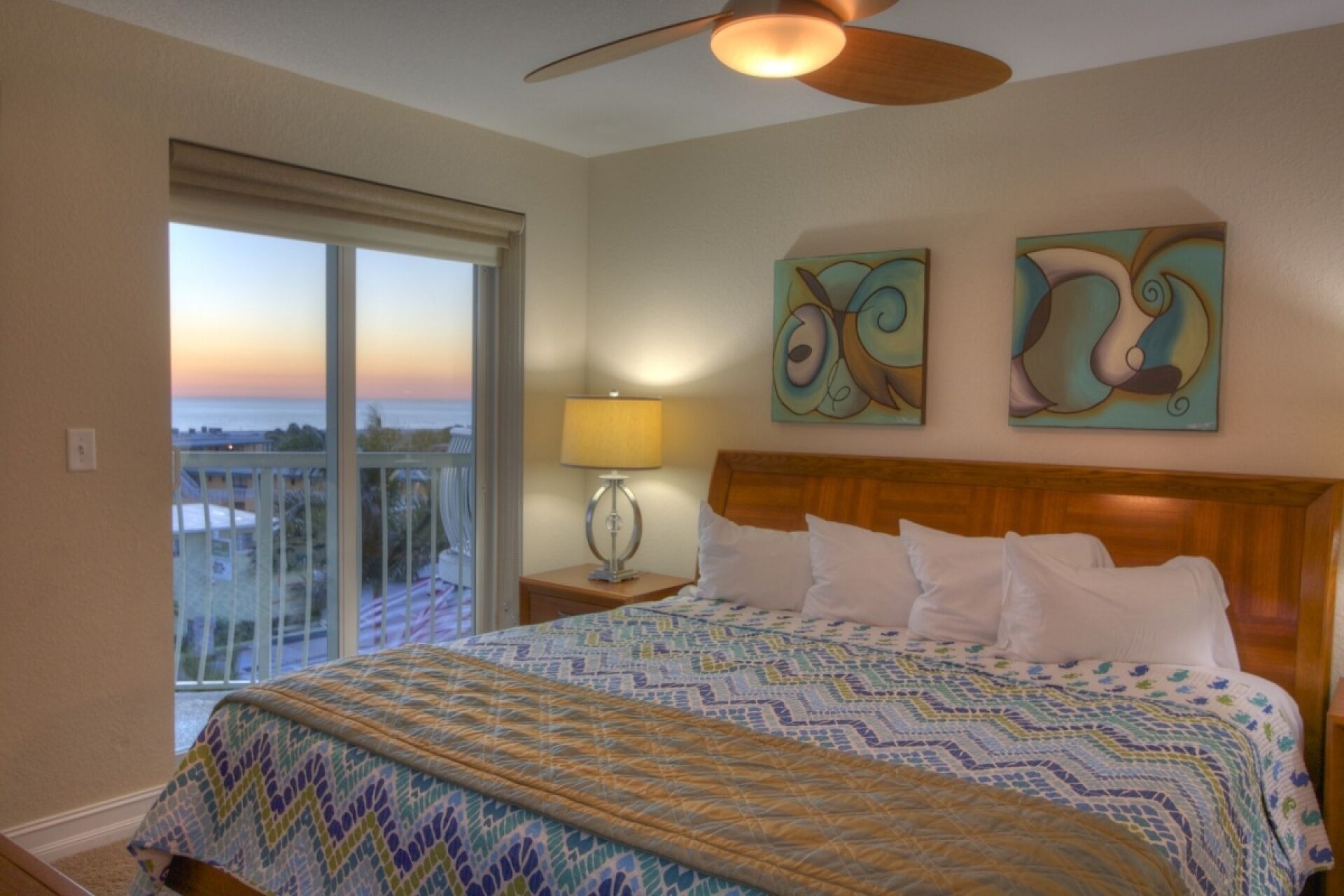 A hotel room with a large bed, a ceiling fan, a view of the ocean through a balcony, and abstract art on the wall.