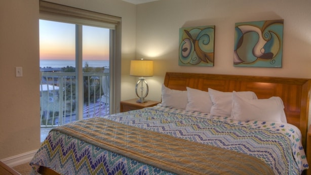 A hotel room with a large bed, a ceiling fan, a view of the ocean through a balcony, and abstract art on the wall. - Gulf Views from Balcony. Heated Pool. Beautiful Property Directly Across from the Beach! (Treasure Island)