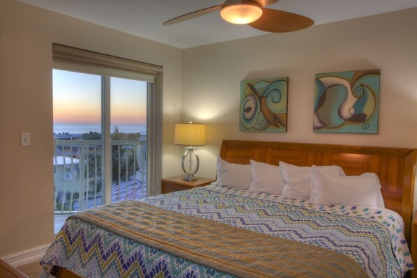 A hotel room with a large bed, a ceiling fan, a view of the ocean through a balcony, and abstract art on the wall. - Gulf Views from Balcony. Heated Pool. Beautiful Property Directly Across from the Beach! (Treasure Island)