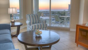 A room with a view of the ocean at sunset, a striped armchair, a round table with a bowl of fruit, and a blue sofa. - Gulf Views from Balcony. Heated Pool. Beautiful Property Directly Across from the Beach! (Treasure Island)