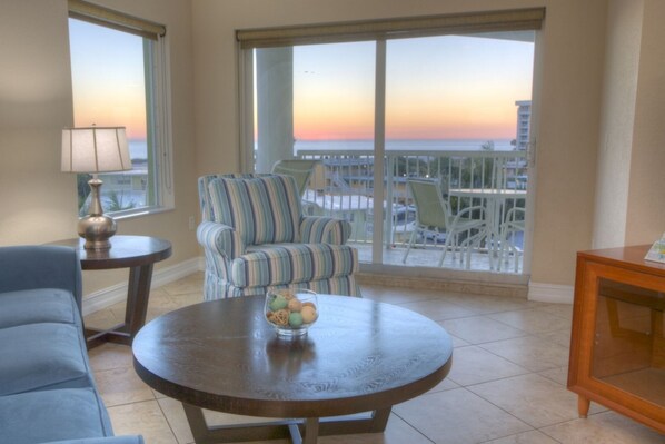 A room with a view of the ocean at sunset, a striped armchair, a round table with a bowl of fruit, and a blue sofa. - Gulf Views from Balcony. Heated Pool. Beautiful Property Directly Across from the Beach! (Treasure Island)