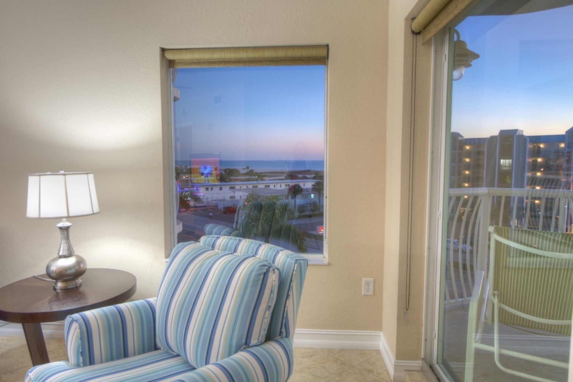 Ein Zimmer mit einem gestreiften Sessel, einem kleinen Tisch mit Lampe und einem großen Fenster mit Blick auf Strand und Häuser.
