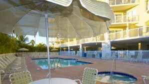 A pool area with lounge chairs and umbrellas, surrounded by a multi-level building with balconies. - Gulf Views from Balcony. Heated Pool. Beautiful Property Directly Across from the Beach! (Treasure Island)