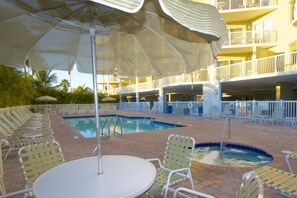 A pool area with lounge chairs and umbrellas, surrounded by a multi-level building with balconies. - Gulf Views from Balcony. Heated Pool. Beautiful Property Directly Across from the Beach! (Treasure Island)