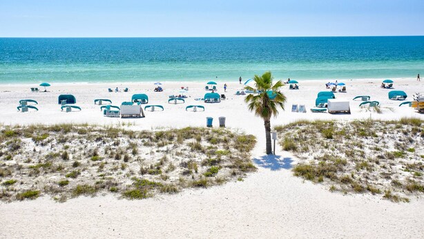 A beach with white sand, clear blue water, and several blue umbrellas and lounge chairs.