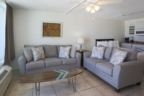 A living room with two grey sofas, a wooden coffee table, a painting on the wall, a ceiling fan, and a kitchenette in the background.