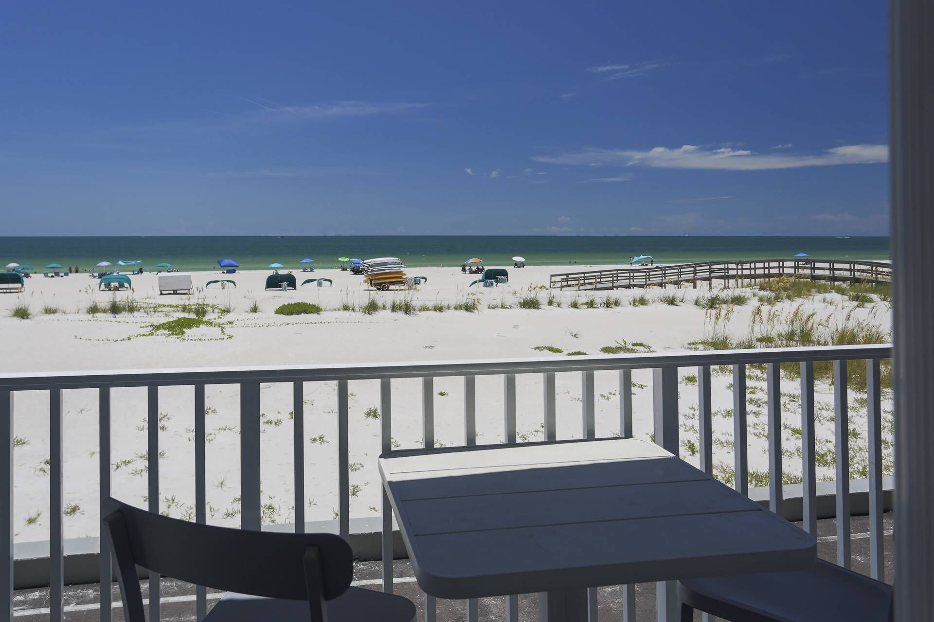 A beach view from a balcony with a table and chair, a wooden deck, and beach umbrellas.