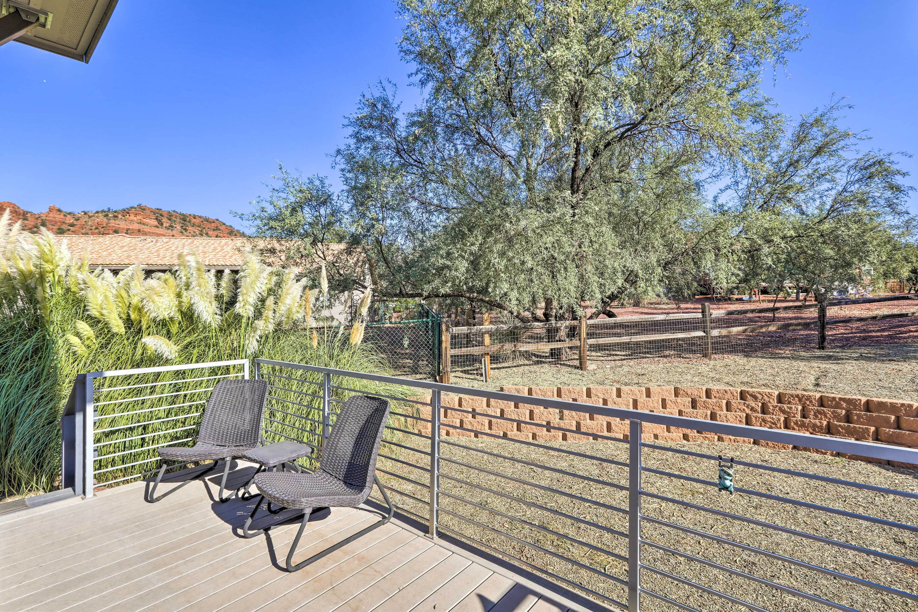 A balcony with two wicker chairs, a view of a fenced area with trees and a distant red hill.