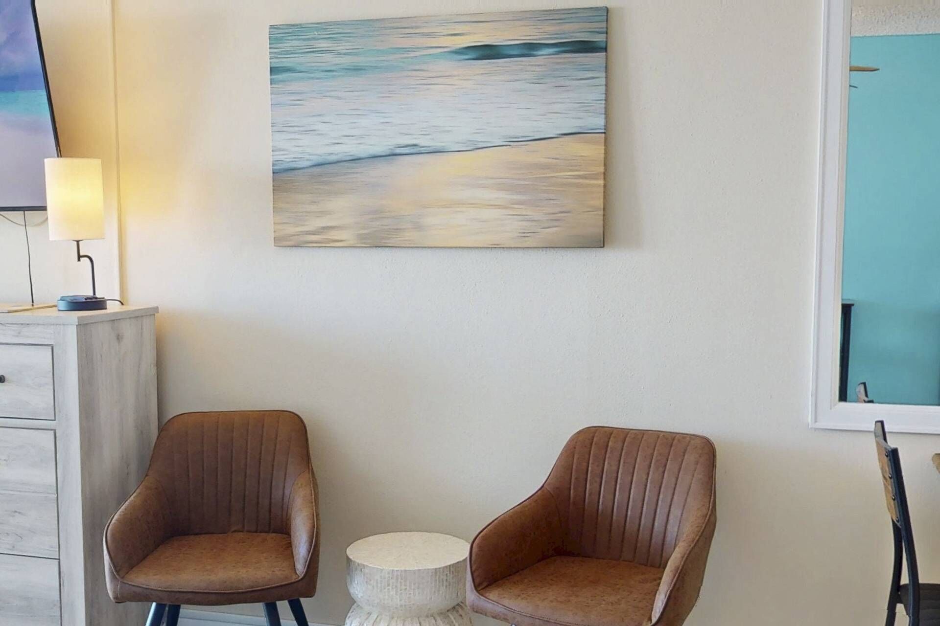 A room with two brown upholstered chairs, a small round table, and a painting of a beach scene on the wall.