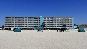 A large, multi-story hotel with numerous balconies, a sandy beach in front, and a clear blue sky.