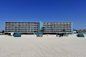 Un hotel grande de varios pisos con numerosos balcones, una playa arenosa frente al establecimiento y un cielo azul despejado.