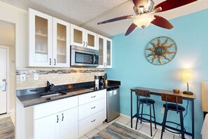 Una cocina compacta con gabinetes blancos, microondas y cafetera. Hay un ventilador de techo, un adorno mural con motivo de sol, y una zona de bar con taburetes.