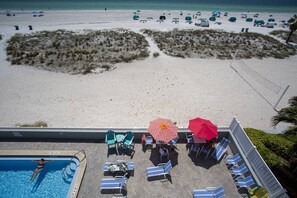 A pool area with lounge chairs and umbrellas, overlooking a beach with umbrellas and people.