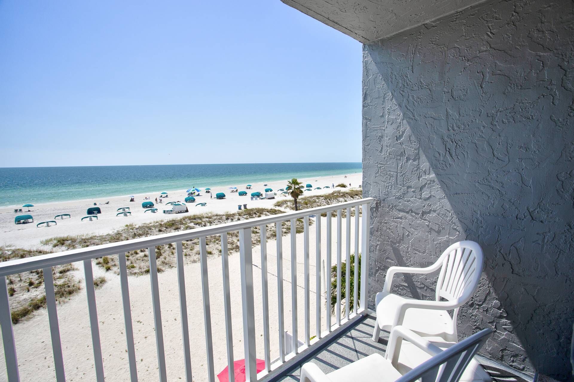 A balcony with white chairs overlooking a beach with umbrellas and people.