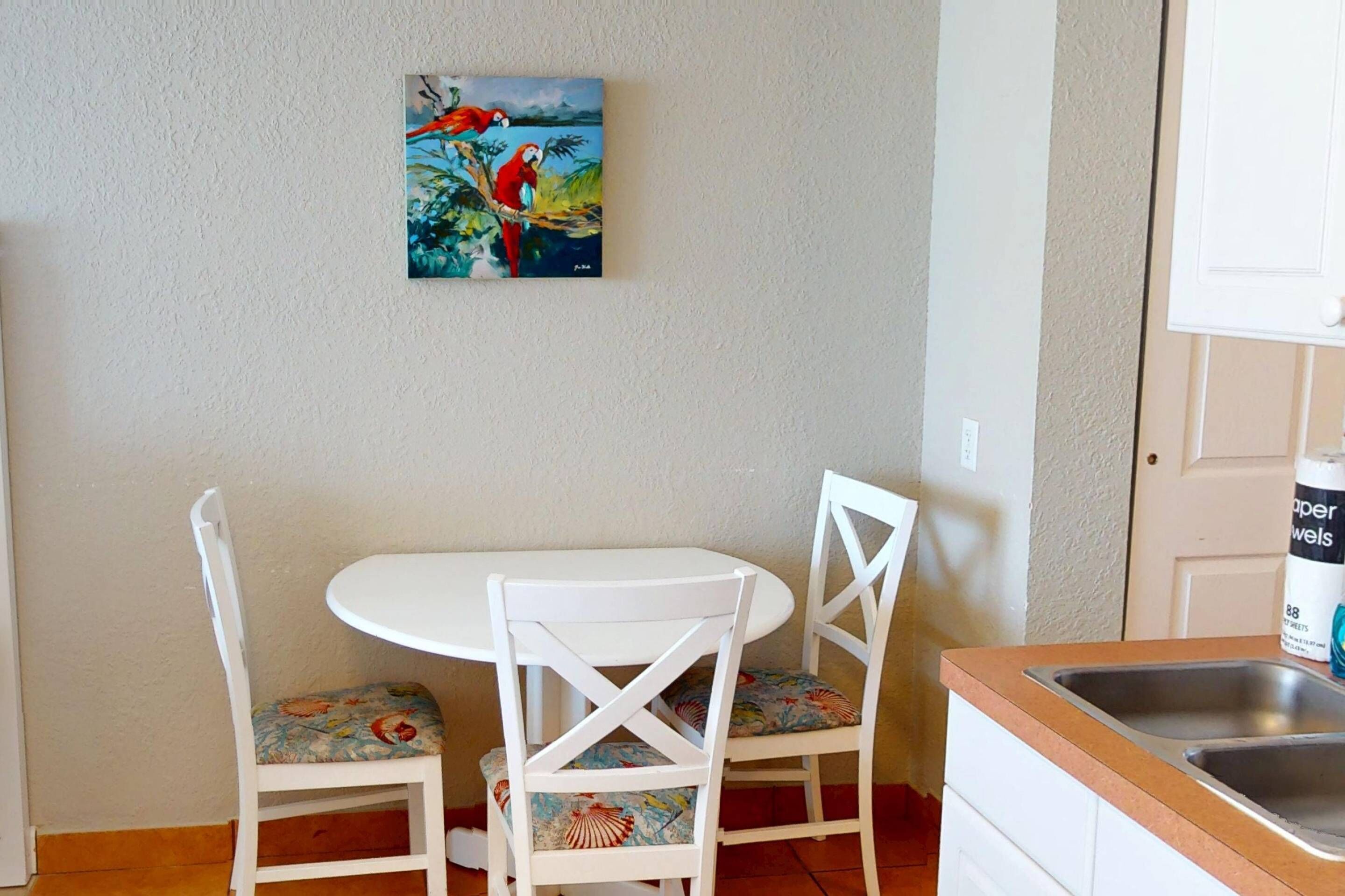Un petit coin repas avec une table blanche et deux chaises blanches ornées de coussins à motifs floraux. Un tableau représentant des perroquets est accroché au mur, au-dessus de la table.