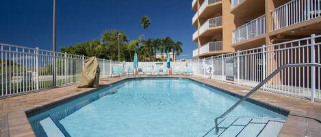 A swimming pool with a tiled deck, surrounded by a white fence, and adjacent to a multi-story building with balconies.
