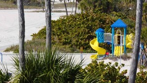 Un parque infantil con un tobogán amarillo y una estructura azul para escalar, rodeado de follaje y una zona de arena.
