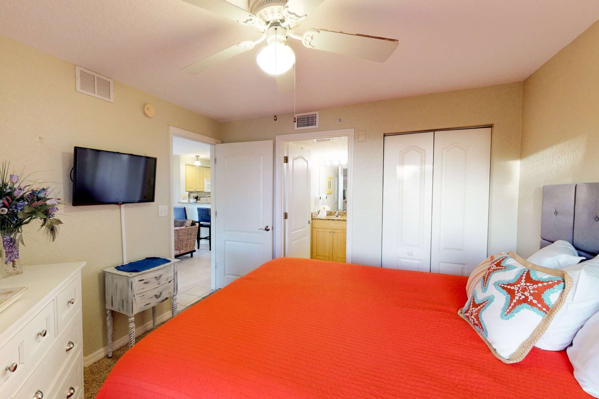 A bedroom with a large bed, a ceiling fan, a television, and a view into the kitchen.