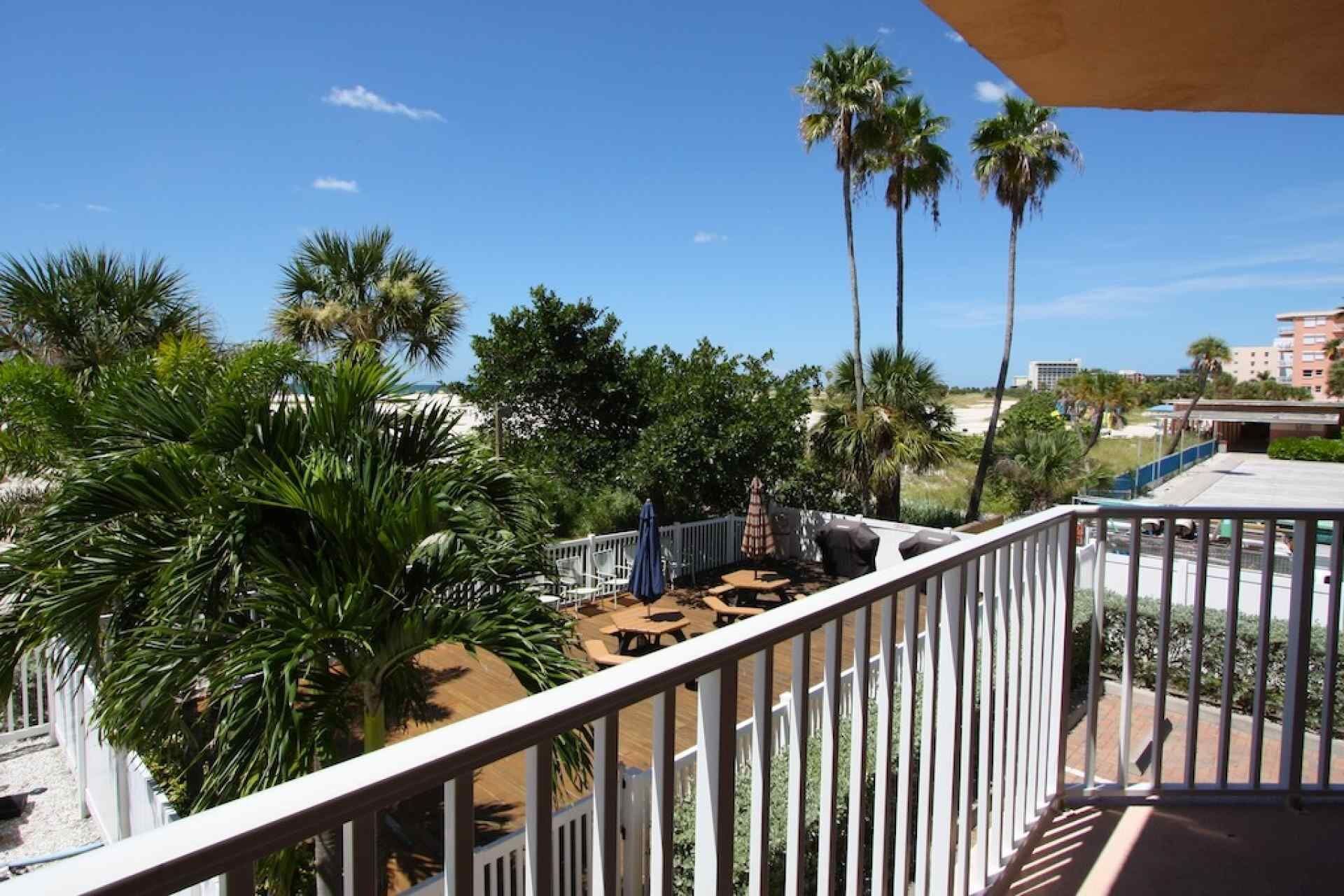 Ein Balkon mit Blick auf Palmen und eine Sandfläche, ein weißes Geländer sowie ein Gebäude im Hintergrund.