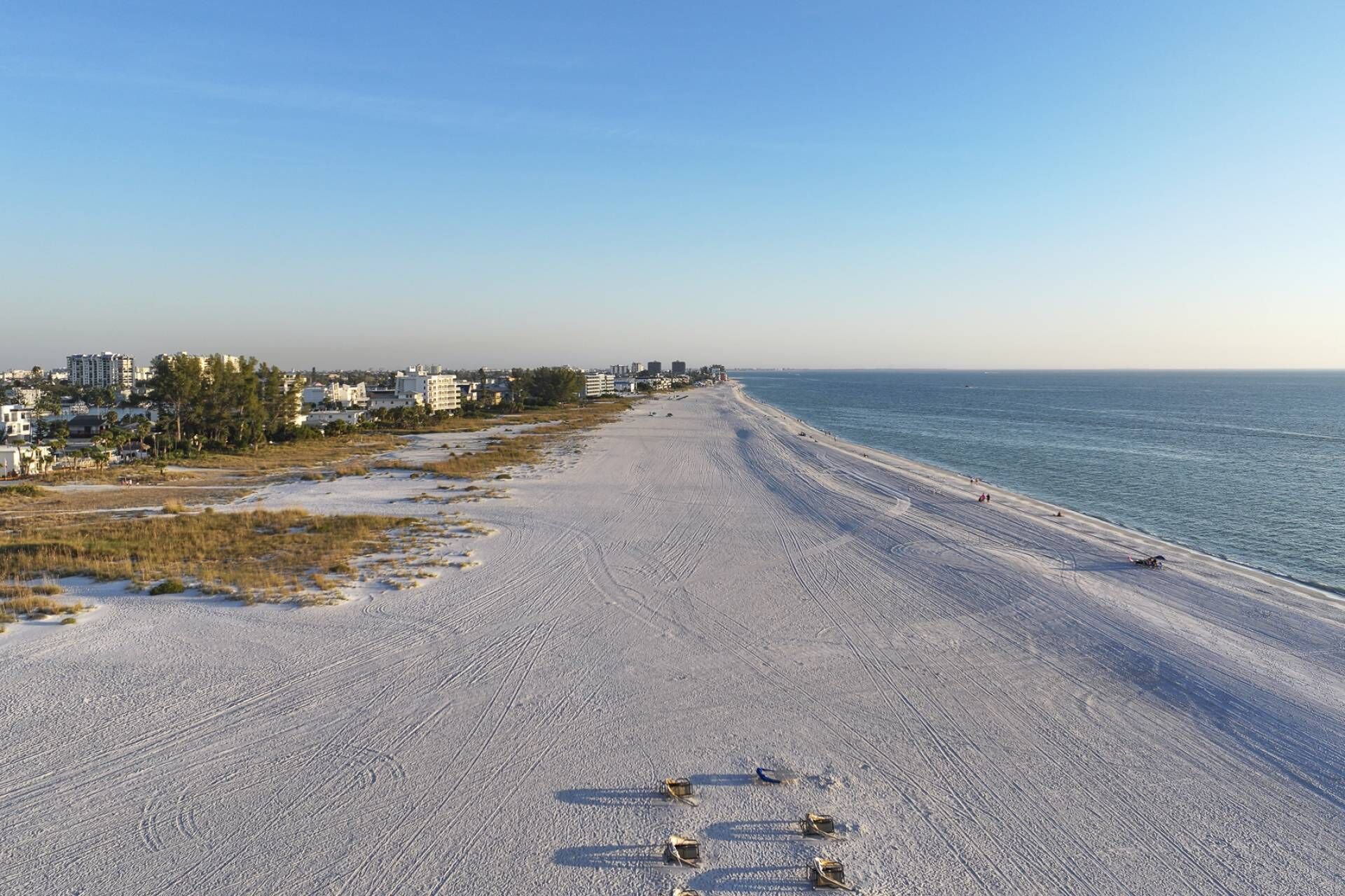 Ein weiter Sandstrand, klarer Himmel, einige Gebäude im Hintergrund und Personen, die am Ufer entlanggehen.