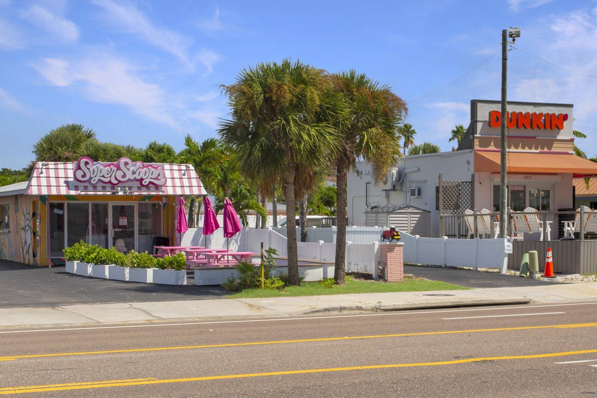 A Super SoCoos store with a pink and white striped awning, a Dunkin' Donuts across the street, and a palm tree in the foreground.