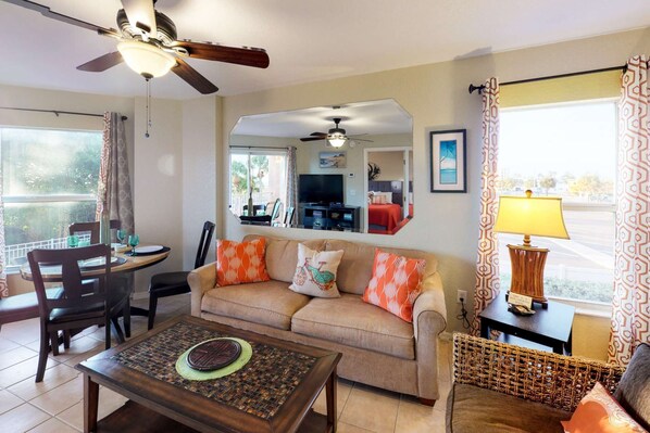 A living room with a sofa, coffee table, and a TV. There is a dining area with chairs and a table. The room has a ceiling fan and a window with curtains.