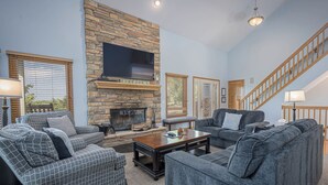 A living room with a stone fireplace, a flat-screen TV, and a staircase. - Outdoor Hot Tub, Pool Table, Huge Fire Pit - Close 2 Pools! (Galena)