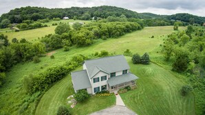 A two-story house with a green roof, surrounded by lush green fields and trees.