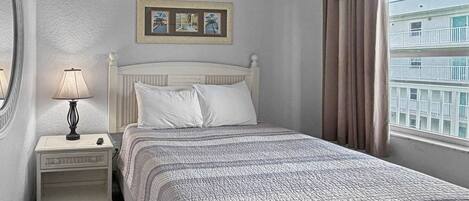 Uma cama bem arrumada com colcha estampada, uma luminária de cabeceira, mesa de cabeceira e um quadro emoldurado acima da cama.