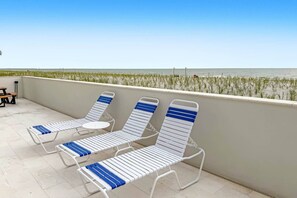 A balcony with three lounge chairs and a small table, overlooking a beach with grass and the sea.
