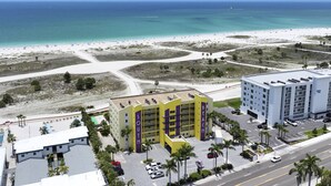 A hotel with a central building featuring a yellow facade and purple accents, adjacent to a beach with clear blue waters and sandy shores. - Modern 2Br/2Ba Beachfront Condo with an Incredible View. Amenities, Pool, Beautiful Beach (Treasure Island)