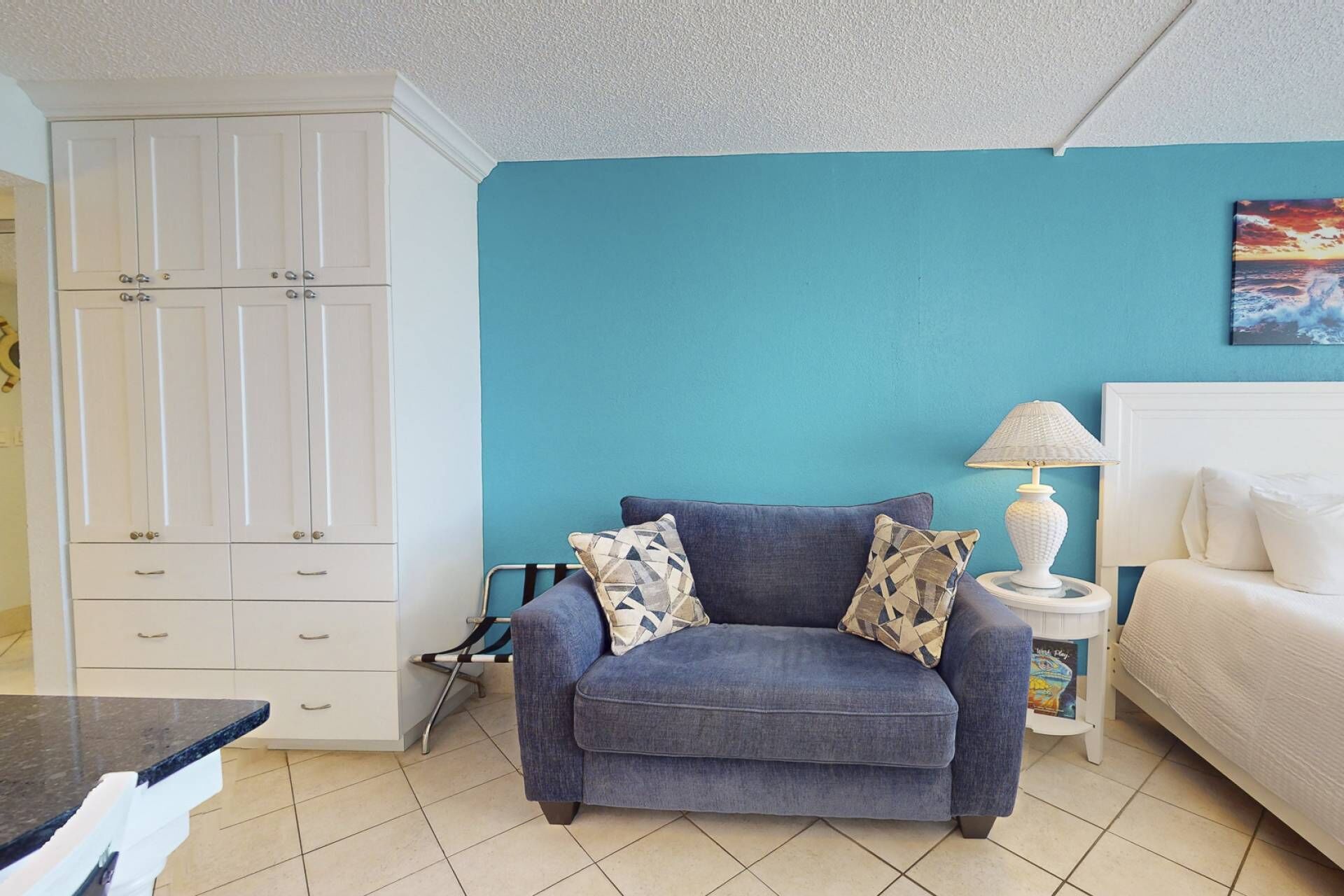 A room with a blue wall, a grey sofa with patterned cushions, a white bedside table with a lamp, and a white wardrobe with multiple doors.