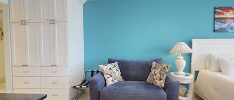 A room with a blue wall, a grey sofa with patterned cushions, a white bedside table with a lamp, and a white wardrobe with multiple doors.