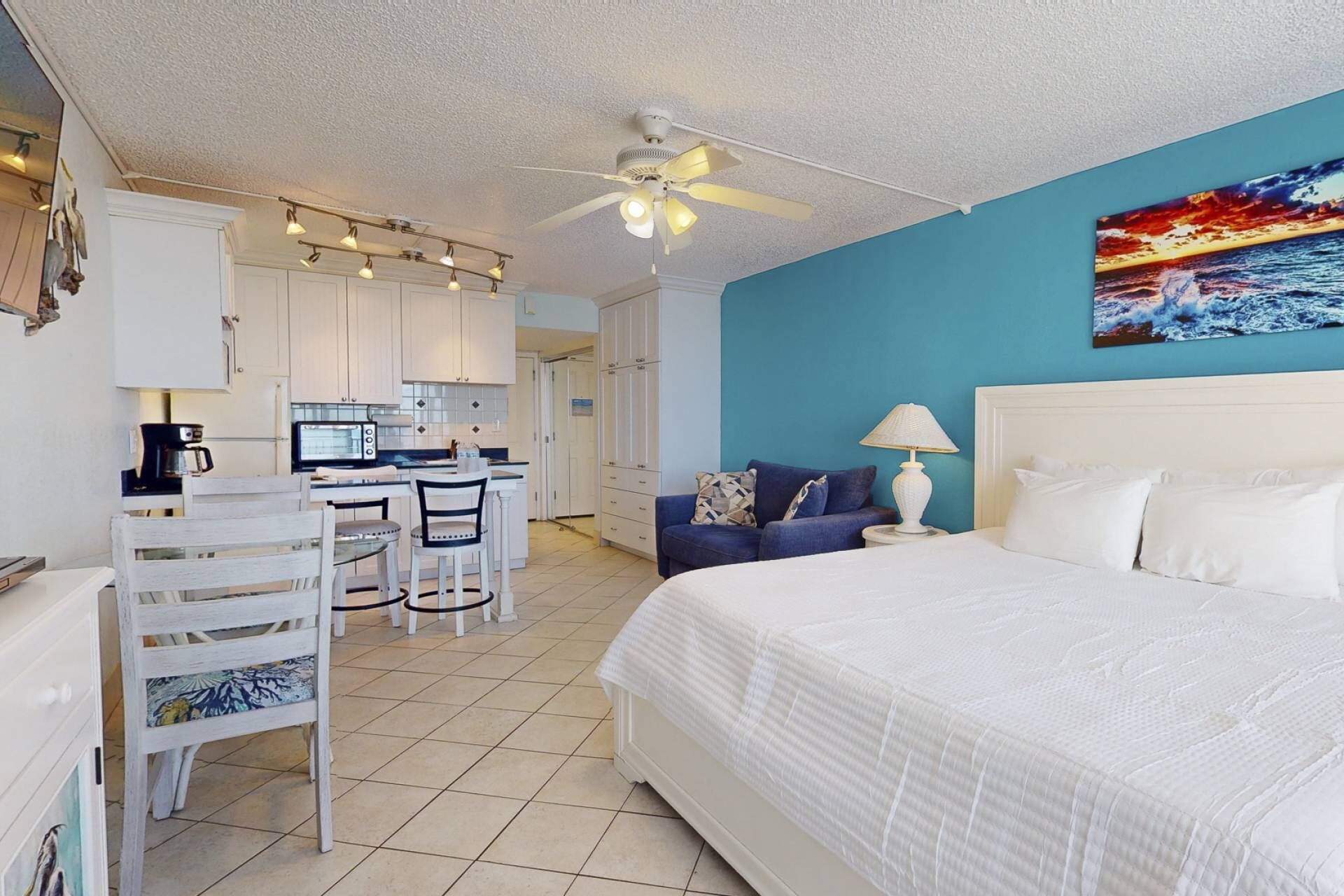 A hotel room with a bed, a desk with chairs, a kitchenette, and a ceiling fan.