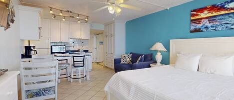 A hotel room with a bed, a desk with chairs, a kitchenette, and a ceiling fan.